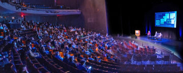 Panoramic view of auditorium with SEJ Conference audience members watching 3 figures seated on stage (Jamieson, Mann, and Weiss).