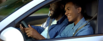 Teenage boy behind the wheel of a car with an adult man in the passenger seat.