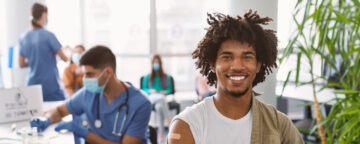 Black man with band-aid from vaccination on upper arm smiles at camera, with medical providers wearing PPE in the background.