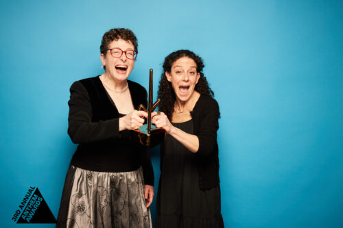 2 women in semi-formal black dresses hold up a bronze trophy from the Anthem Awards.