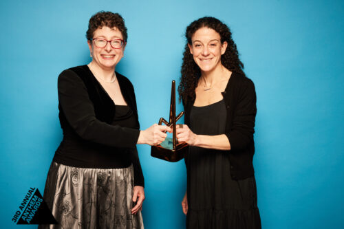 2 women in semi-formal black dresses hold up a bronze trophy from the Anthem Awards.