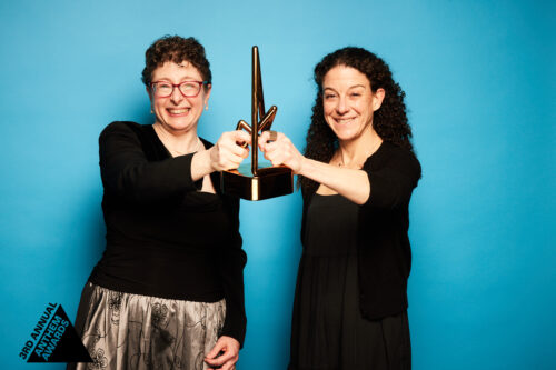 2 women in semi-formal black dresses hold up a bronze trophy from the Anthem Awards.