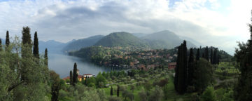 Scenic view with body of water on left, green trees in foreground, old-style terracotta-roofed houses in the middle-ground, and green hills in the distance.