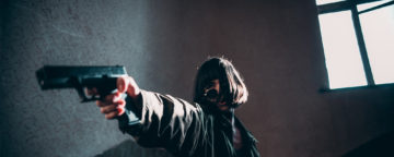 Person in oversized coat, sunglasses, and hair falling across their face pointing a gun. Credit: Elijah O'Donnell.