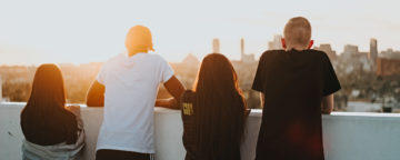 4 teens stand with their backs to the camera looking out towards a sunny city skyline. Slider art for study on 13 Reasons Why and adolescent suicide.